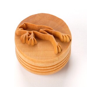 May include: A light brown, wooden stamp with a carved frog design. The stamp is round with a layered base and a detailed frog carving on top. The frog is depicted with outstretched limbs.