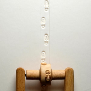 Op de afbeelding: Een houten stempel met een schoenafdrukontwerp, waarmee een spoor van afdrukken op een wit oppervlak wordt gecreëerd. De stempel heeft een houten handvat en is gemaakt van lichtgekleurd hout.