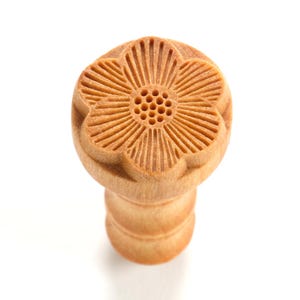 May include: A wooden flower-shaped stamp with a detailed design and a small hole in the center. The stamp is light brown in color and has a cylindrical base.