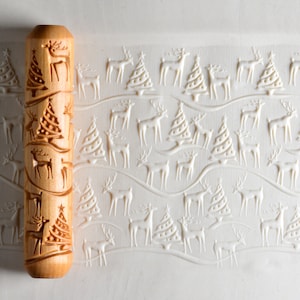 May include: A wooden rolling pin with a repeating pattern of stylised reindeer and Christmas trees. The rolling pin is on a white surface with an impression of the rolling pin's design.