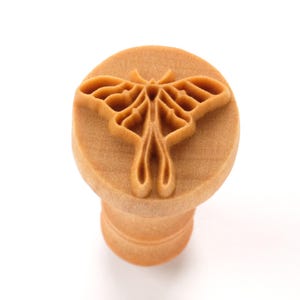 May include: A wooden stamp featuring a moth design. The stamp has a circular top with the moth carving and a cylindrical handle. The moth has detailed wing patterns and a delicate body. The stamp is made of light brown wood.