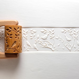 Könnte beinhalten: Ein hellbrauner Nudelholz mit einem geschnitzten Design von Vögeln und Blättern. Ein weißes geprägtes Muster von Vögeln und Zweigen ist ebenfalls zu sehen, das eine dekorative Bordüre bildet.