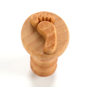 May include: A wooden stamp with a footprint design. The stamp is round and has a small handle.