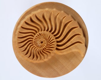 May include: A wooden stamp with a detailed carving of a nautilus shell. The shell is carved in a light brown color and has a spiral pattern.