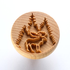 May include: A wooden round stamp with a carved design of a moose standing in a forest. The moose is facing the left side of the stamp and has large antlers. The trees are on either side of the moose and have a detailed design.