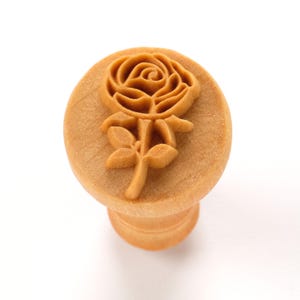 May include: A wooden rose-shaped cookie stamp. The stamp features a detailed floral design of a rose blossom and stem. The stamp is light brown and has a round handle.