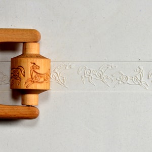 May include: A wooden rolling pin with a carved horse design. The rolling pin is light brown and features a cylindrical center with a repeating pattern of horses. The horses are also imprinted on the light-colored surface.