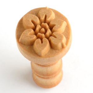 May include: A light brown wooden stamp featuring a circular head with a carved floral pattern. The design consists of five symmetrical, heart-shaped petals arranged around a central point, set atop a cylindrical handle with a smooth finish.