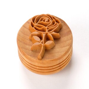 Könnte beinhalten: Ein Holzstempel mit einem geschnitzten Rosenmotiv. Der Stempel ist kreisförmig mit einem geschichteten Sockel und einer detaillierten Blumenschnitzerei auf der Oberseite. Das Holz hat eine hellbraune Farbe und sichtbare Maserung.