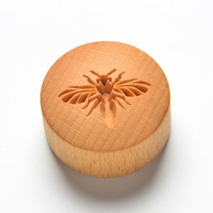 May include: A round wooden cookie cutter featuring a carved bee design. The cutter is made from light brown wood and has a smooth, polished finish.