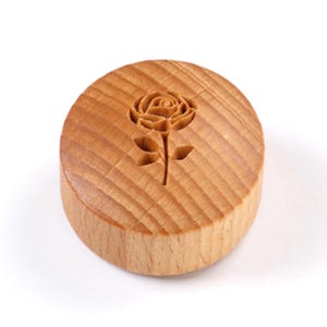 Könnte beinhalten: Ein runder Holzstempel mit einem geschnitzten Rosenmotiv. Der Stempel besteht aus hellem Holz und weist ein detailliertes Blumenmuster auf. Die Rose ist mittig auf dem Stempel platziert, mit Stiel und Blättern. Die Holzmaserung ist sichtbar.