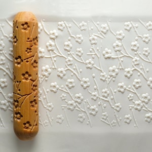 Puede incluir: Un rodillo de madera con un diseño tallado de flores de cerezo. El rodillo está sobre una superficie blanca con un diseño en relieve similar.