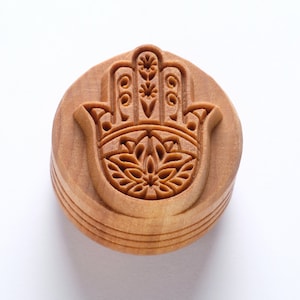 Pode incluir: Um carimbo de madeira em forma de mão de Hamsa com entalhes intrincados. A mão de Hamsa é um símbolo de proteção e boa sorte.
