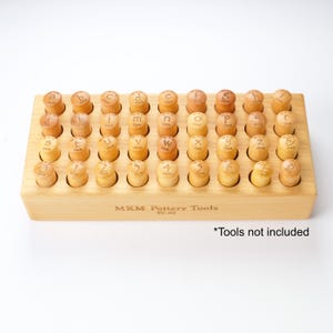 May include: A wooden pottery tool set with a light wood base and individual stamps for letters and numbers. The stamps are arranged in a grid, with the text "MKM Pottery Tools TC-02" on the base. The text "Tools not included" is also present.