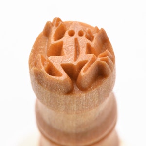 May include: A wooden stamp with a carved frog design. The stamp is light brown and has a smooth surface.