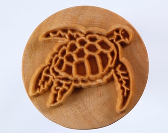 May include: A wooden round stamp with a detailed carving of a sea turtle. The turtle is facing right and has a textured shell.