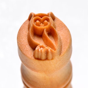 May include: A close-up of a wooden stamp featuring a carved owl design. The stamp is light brown and has a cylindrical shape with the owl's details carved into the top surface. The owl has large eyes and a small beak.