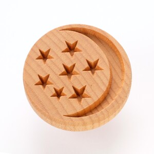 May include: A circular wooden coaster featuring a crescent moon design and seven star-shaped cutouts. The coaster is crafted from light-toned wood with a natural finish. The stars are uniformly arranged across the surface.