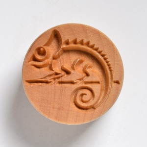 May include: A wooden rolling pin with a carved chameleon design. The chameleon is facing right and has a curled tail. The rolling pin is made of light brown wood.