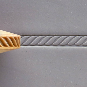 May include: A close-up of a wooden stamp with a rope design. The stamp is being used to create an embossed rope design on a grey surface.