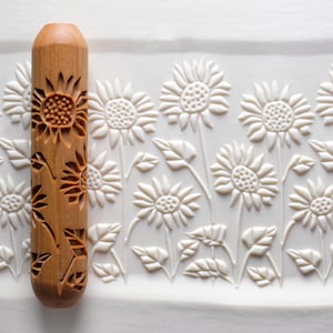 May include: A wooden rolling pin with a sunflower design is shown next to a white surface with a sunflower pattern. The rolling pin is light brown and features carved sunflowers and leaves. The white surface has a repeating sunflower pattern.