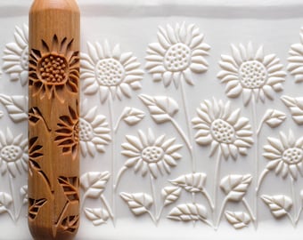 May include: A wooden rolling pin with a sunflower design is shown next to a white surface with a sunflower pattern. The rolling pin is light brown and features carved sunflowers and leaves. The white surface has a repeating sunflower pattern.