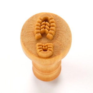 May include: A wooden stamp with a footprint design. The stamp has a round top with the footprint impression and a cylindrical handle. The wood is a light brown color, and the design is carved into the wood.