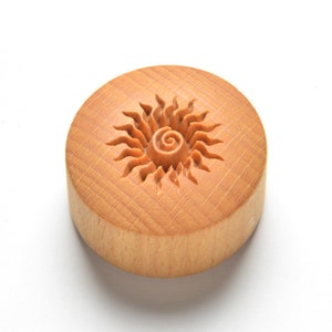 May include: A round wooden stamp with a carved sun design. The sun has a spiral in the center and rays extending outward.