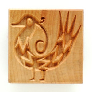 May include: A square, wooden cookie stamp featuring a carved bird design. The bird has a curved beak, a spiral body, and a tail with pointed edges. The stamp is made of light-colored wood.