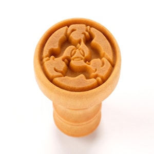 May include: A light brown bottle stopper with a detailed, circular design. The top features a raised relief of a stylized figure. The stopper has a cylindrical base. The object is set against a white background.