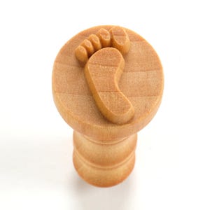 May include: A wooden stamp featuring a footprint design. The stamp is light brown and has a round top with a small cylindrical base.