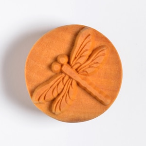 May include: A round, orange-brown wooden button with a detailed dragonfly design carved into the surface.