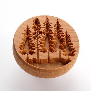 May include: A circular light brown wooden stamp featuring a deeply carved relief design of a cluster of four coniferous trees. The wood grain is visible across the smooth, rounded surface of the stamp, highlighting the detailed texture of the tree branches and trunks.