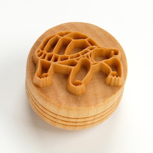 Op de afbeelding: Ronde houten stempel met een schildpadontwerp. De stempel is lichtbruin en heeft een gelaagde basis. De omtrek van de schildpad is verhoogd, met schild, poten en kop. Diameter ongeveer 6 cm.