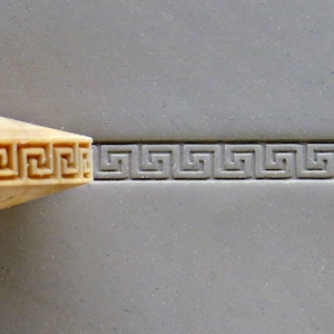 May include: A close-up of a wooden stamp with a Greek key design being used to create an imprint on a grey surface.