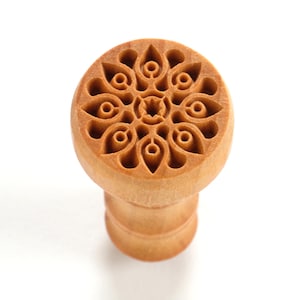 May include: A wooden stamp with a detailed carved design of a circular pattern with multiple petals. The stamp is light brown and has a cylindrical handle.