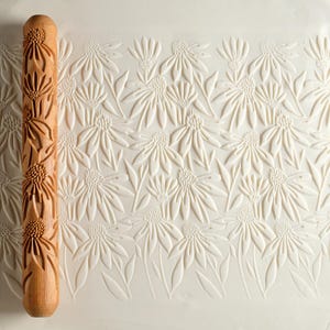 Könnte beinhalten: Ein Nudelholz aus Holz mit einem floralen Muster. Das Nudelholz wird verwendet, um ein geprägtes Muster auf Teig oder Fondant zu erstellen. Das Muster zeigt ein sich wiederholendes Design aus stilisierten Blumen.
