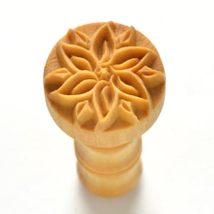 May include: A wooden stamp with a carved floral design. The stamp is light brown and has a round top with a cylindrical base.