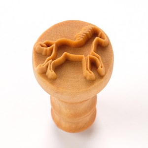 May include: A light brown, wooden stamp with a horse design. The horse is in profile, with a detailed mane and tail. The stamp has a round top and a cylindrical handle.