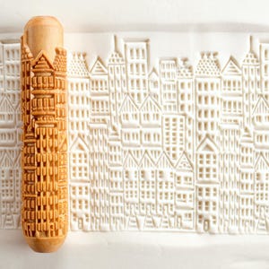 May include: A wooden rolling pin with a detailed cityscape design. The pin is light brown and features a repeating pattern of buildings. The impression on the dough shows a detailed city skyline, perfect for baking themed cookies or pastries.
