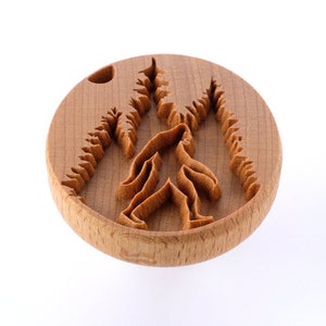 May include: A wooden round cookie cutter with a carved design of Bigfoot walking through a forest.