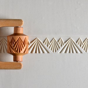May include: A wooden rolling pin with a carved design of a repeating fan pattern. The rolling pin is being used to create a decorative border on a white surface.