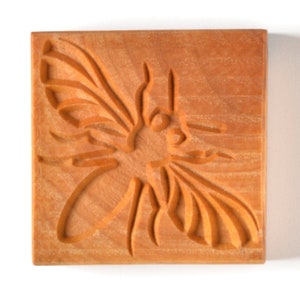 May include: A square, wooden block with a carved design of a bee. The bee's wings and body are detailed with lines and curves. The wood has a warm, orange-brown color and a visible grain.