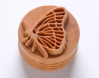 May include: A wooden round ornament with a carved butterfly design. The butterfly is detailed with intricate wing patterns and is a light brown color.