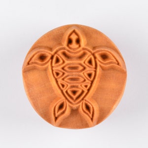 May include: A round, orange-brown clay stamp with a detailed design of a turtle. The turtle has a patterned shell and a simple face.
