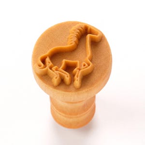 May include: A wooden stamp with a horse design. The stamp features a circular top with a raised outline of a horse and a cylindrical handle. The wood has a light brown color, and the design is carved into the top surface.