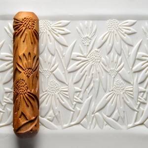 May include: A wooden rolling pin with a carved design of flowers and leaves. The rolling pin is on a white surface with a similar design.