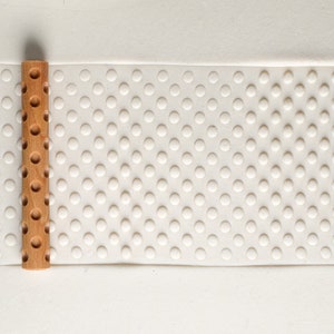 May include: A wooden rolling pin with a pattern of circles on the surface. The rolling pin is on a white surface with a pattern of circles.