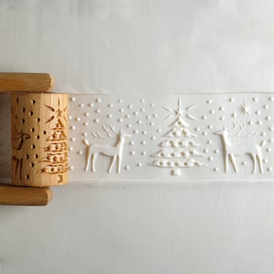 May include: A wooden rolling pin with a carved Christmas tree and deer design. The pin is next to a white embossed pattern of deer, a Christmas tree, snowflakes, and stars. The rolling pin has wooden handles.