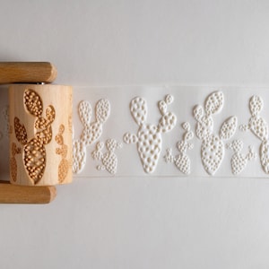 Peut inclure: Un rouleau à pâtisserie en bois avec un motif de cactus. Le rouleau à pâtisserie est utilisé pour créer des motifs en relief sur le fondant ou l'argile. Le rouleau à pâtisserie est présenté avec une surface blanche sur laquelle est embossé un motif de cactus.